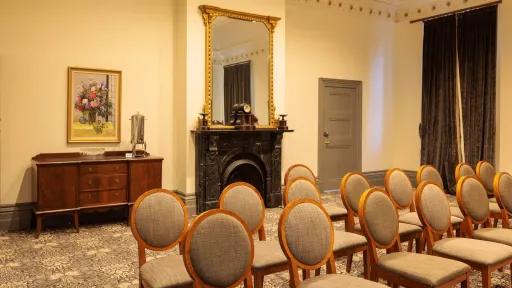 A room arranged for a presentation, with rows of chairs facing forward. The room has a buffet table, floral painting, a coffee urn, a black marble fireplace, a mirror. A doorway is to the right of the fireplace. The room has decorative molding around the ceiling and patterned carpet.