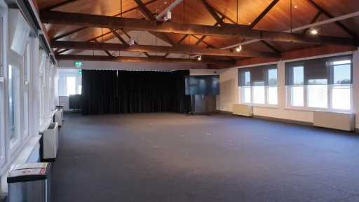 Wide shot of an empty event space with a high, angled wooden ceiling supported by exposed beams. Dark gray carpet covers the floor. A wall of windows is on the left, and another wall of windows with dark blinds is on the right. Black curtains are draped on the far wall, with a large flat-screen TV in front.