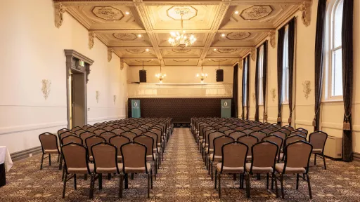 An interior view of a large room set up for an event. Rows of chairs face the back of the room, where a raised platform is centered.  The ceiling is ornately decorated with square recessed panels and gold trim and chandeliers  Tall windows line one side of the room, draped with dark curtains. The floor is covered with a patterned carpet.
