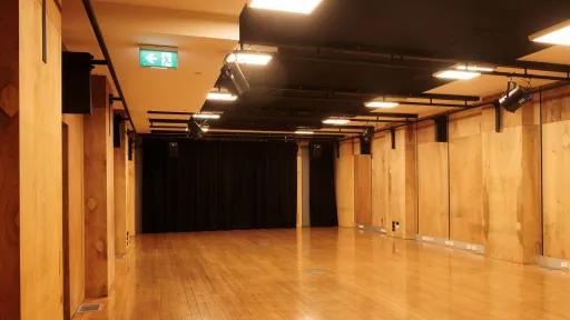 A dance studio with a wood floor and wood panel walls. There is a black curtain at the far end of the room. Overhead lights are mounted on a black ceiling. A green exit sign is above the doorway on the left.