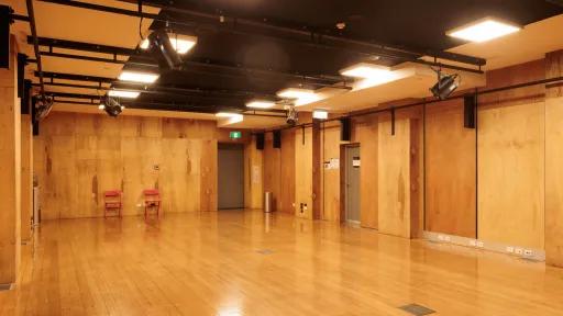 Interior view of a dance studio or theater rehearsal space with wooden floors and walls, overhead lighting and rigging, and two red chairs.