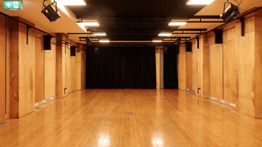 A room with a wooden floor and wood-paneled walls, equipped with lighting and a stage area with black curtains. The room also contains speakers mounted on the walls and an exit sign.