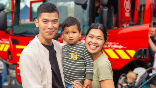 Smiling man and woman stand holding small child with fire engine in background
