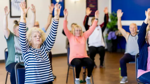 A group of older adults are sitting on chairs with their hands in the air. 