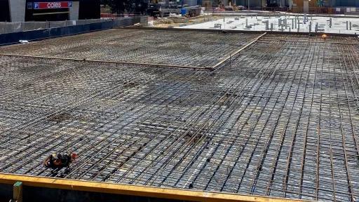 In the foreground, a dense grid of reinforcing steel (rebar) is laid out, ready for concrete pouring. Behind the rebar, there's evidence of ongoing construction activities, including concrete foundations, exposed framework, and equipment. In the background, there's a larger structure under construction, with its concrete walls partially completed.