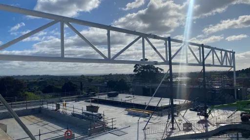 A construction site under a bright sky with scattered clouds. A large metal beam structure is in the foreground, and unfinished concrete structures, safety rails, and equipment are visible in the midground. Distant trees and hills complete the background.