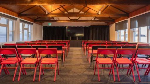 A room with rows of folding chairs facing a stage with a large screen and black curtains. The room has a high, wooden ceiling with exposed beams and several windows on both sides. The floor is carpeted.