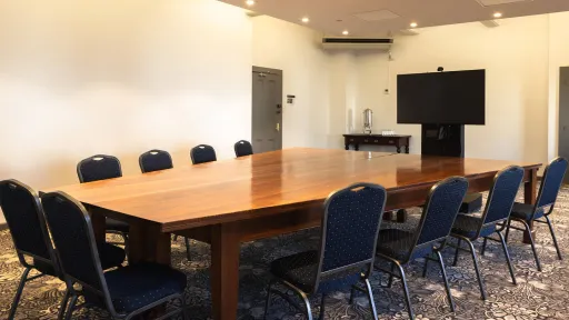 A long rectangular wooden table is surrounded by blue patterned chairs in the meeting room. A large screen is mounted on the wall at one end of the table.