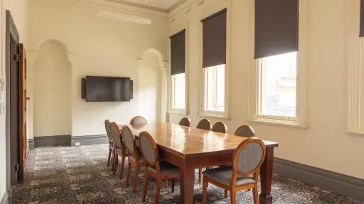 A meeting room featuring a long rectangular wooden table with 10 chairs. A TV sits on the far wall. There are tall windows and patterned carpet.