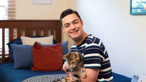 A young person is sitting on a single bed. They are smiling at the camera and holding a cat.