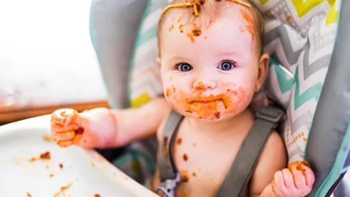 A baby sitting in a highchair, face, hands and highchair tray covered in a red sauce.