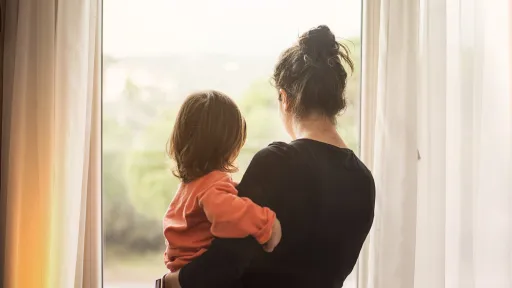 A woman holding a small child looks out the window.