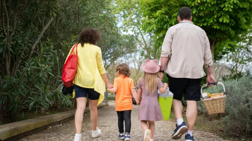 Two adults and 2 children are walking with backs away from camera in a park. The adults are holding picnic bags.