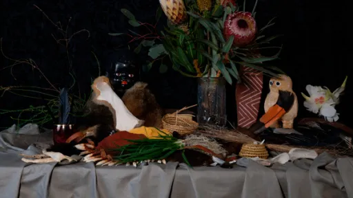 A vase with flowers sits on a grey tablecloth. In front of the vase is a cluster of plants, shells, reed baskets and dolls. 