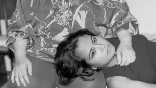 A black and white photo portrait of a young person lying with their head on an older person’s lap. The older person sits upright, hands resting on their legs, while the younger person looks directly at the camera.
