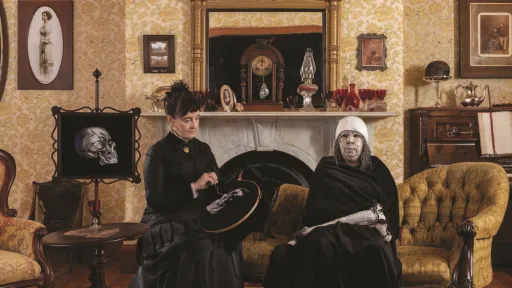 A staged Victorian-style interior with patterned wallpaper and antique furniture, where two women (artists megan evans and Maree Clarke) dressed in black sit together on an upholstered chair.