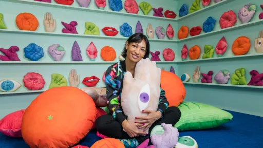 Artist Kate Beynon sits on a blue carpet in front of light teal shelf with colourful soft sculptures of body parts and other motifs. Some have Chinese characters written on them. Kate sits amongst larger soft sculptures and holds one in her arms, which appears to be the back of a hand with an eye on it. Kate is smiling at the camera. 