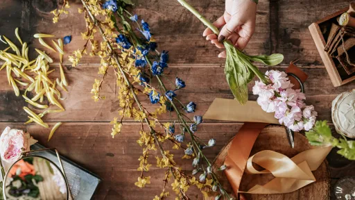 A wooden table scattered with floral arrangement materials, including long stems of yellow and blue flowers, pink blossoms held by a hand, and green leaves. Surrounding the flowers are ribbons in shades of brown and beige, a round wooden board, a small mirror and a box with twine. 