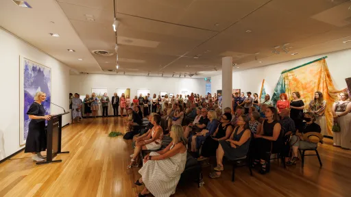 A large crowd is gathered inside an art gallery with wooden floors and white walls covered in contemporary artworks, including a large, blue-toned piece on the left and colourful draped fabric works on the right. A woman stands at a lectern on the left side of the image, speaking to the audience. People are seated in rows at the front while many others stand around the room, filling the space. 