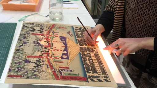 A conservator patches up a torn edge of a yellowed Japanese print using a small paint brush. The print is placed on top of a lightbox. 