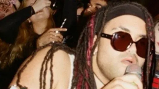 Person with a microphone, wearing sunglasses and has a headband holding back dreadlocks. a dancing crowd is in the background