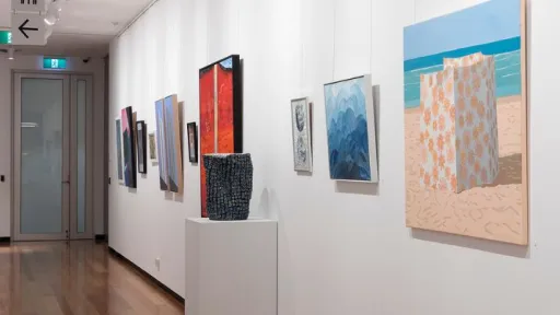 The image shows a long, well-lit gallery space with wooden flooring and white walls adorned with various artworks. The artworks include colourful abstract paintings, geometric compositions, and landscape scenes, all framed or mounted on the walls. The perspective extends down the hallway.