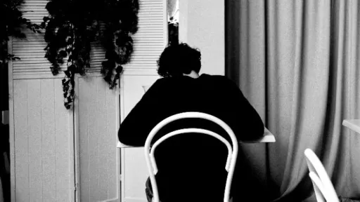 A black-and-white photo of a person seated alone at a table in a cafe. They are facing away from the camera. The space is filled with white chairs and tables, and an arched curtain feature dominates the background. Indoor plants add contrast to the scene.
