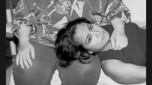 A black and white photo portrait of a young person lying with their head on an older person’s lap. The older person sits upright, hands resting on their legs, while the younger person looks directly at the camera.