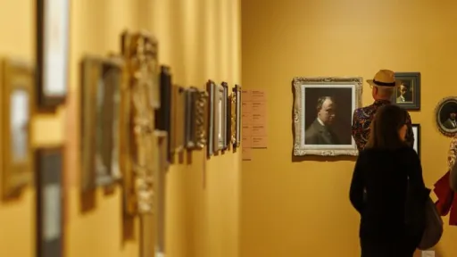 A warmly lit gallery corridor with golden-yellow walls and wooden floors. A long row of small, framed artworks are on the wall to the left, shown in soft focus. At the far end of the space, more framed portraits and small works hang on the wall. Several visitors walk through the gallery, some pausing to look closely at the artwork, while others continue moving. 