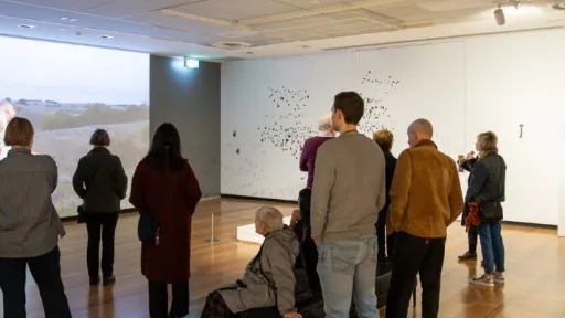 A group of people stand and sit throughout a large gallery with white walls and wooden floors. On the left wall, a large projected video shows an outdoor landscape with a burning object. In the centre, visitors are standing facing the projection and a white wall displaying a delicate installation made of many small black elements suspended in a scattered pattern. Additional framed works hang on the right wall. 