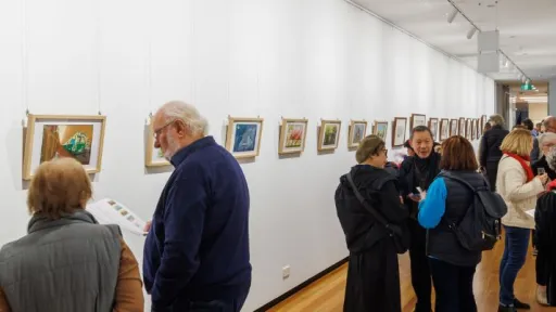 People standing in a long, white-walled gallery corridor with wooden floors, looking at rows of small, framed artworks hung on both sides. Track lighting runs along the ceiling, and visitors gather in small groups throughout the space.