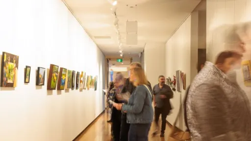 A long gallery corridor with wooden floors and rows of colourful artworks hung on both walls. Several people move through the space, some appearing blurred from motion, while others pause to look at the paintings under bright track lighting.