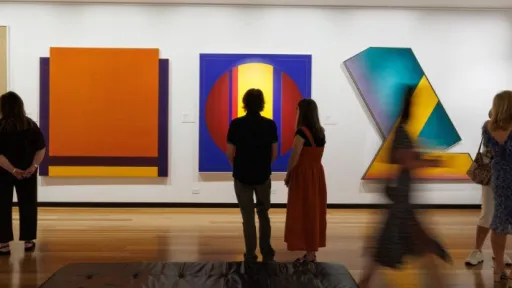 A group of people stand and walk through a gallery space with polished wooden floors. They face a white wall displaying large, bold abstract artworks in vivid colours, including yellow and green, orange, red and blue. Some visitors stand still observing the works, while others move through the scene, appearing slightly blurred. Additional framed artworks hang on the adjacent wall to the right, where more visitors gather in conversation. A black bench sits in the centre foreground.