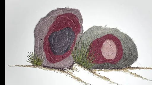 Photo of a mixed media textile artwork on white paper depicting two, rock-like forms. Each form is composed of layered and stitched pieces in muted tones of maroon, grey, black, and purple, creating circular patterns. Between and around the rock shapes, green and brown threads and textured fibres simulate tufts of grass and soil, extending slightly from the surface. 