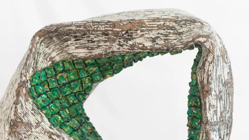 Close-up of a sculptural form with a rough, textured surface in white and brown tones. An arched opening is filled with small, clustered green blocks that resemble carved or moulded cubes. The overall shape curves organically, with etched, scratch-like markings across its surface.