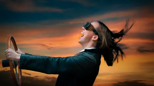 Woman wearing driving goggles and holding a steering wheel has her head thrown back and is shouting with joy.