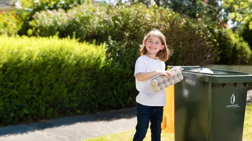 A young girl stands on the nature strip next to a recycling bin with a yellow lid