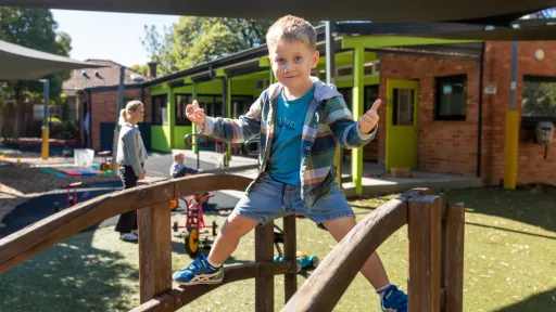 Preschool child stands on wooden bridge in playground giving thumbs-up