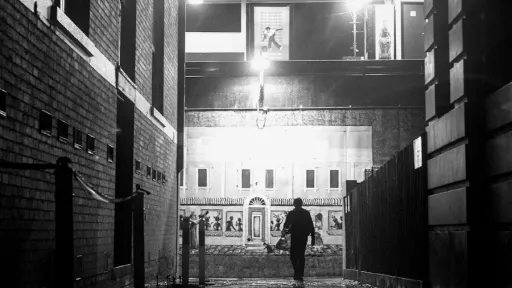 A black and white image of a person walking down a laneway surrounded by buildings
