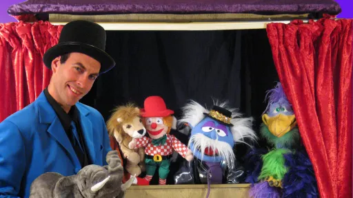 A person wearing a black top hat and blue jacket is standing alongside puppets sitting on a shelf.