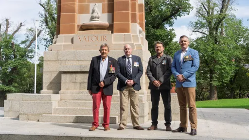 Four men wearing medals pinned to blazers stand in front of war memorial.