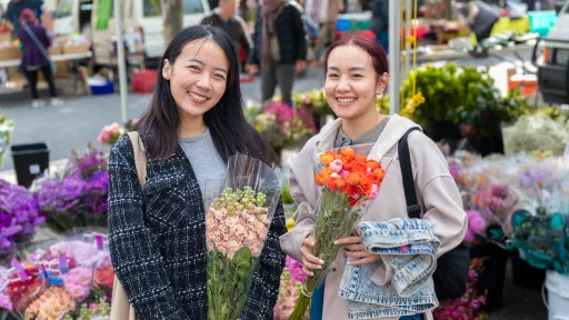 2 people standing in front of a flower stall holding bunches of flowers and smiling.
