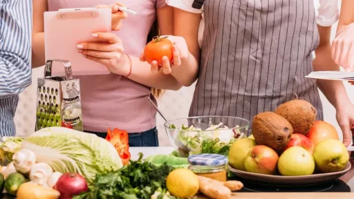 A bench covered in fruit and vegetables. a group of people are looking at a recipe book and taking notes.