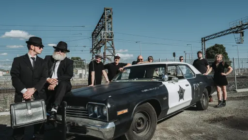 Two men in black suits and hats stand in front of a replica American police car with others in background