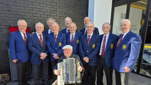 A group of 10 older men standing a one sitting with an accordion. They're wearing blue jackets and red ties.