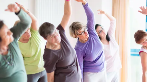 A group of people in loose fitting exercise clothes raise an arm over their heads and lean to the opposite side to stretch.
