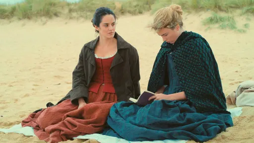 Two females sitting on a beach wearing clothing from the 18th century