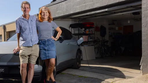 Two people stand smiling in front of grey car outside garage