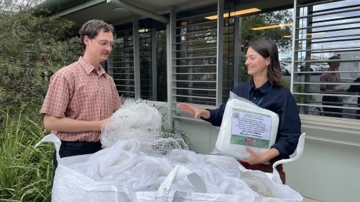 Two people standing with a large bag of bird net. The person on the right holds packaging containing new, legal netting and looks to be explaining something to the person on the left.