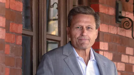 A man stands in front of a brick wall wearing a white shirt and grey jacket